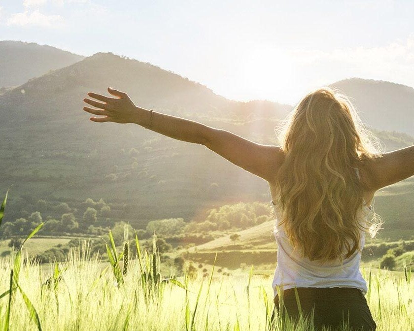 Young woman standing in an open field with arms lifted toward the mountains, expressing freedom, hope, and supernatural joy in God’s presence.