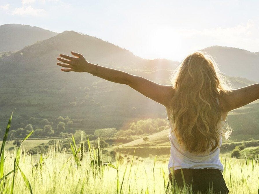 Young woman standing in an open field with arms lifted toward the mountains, expressing freedom, hope, and supernatural joy in God’s presence.