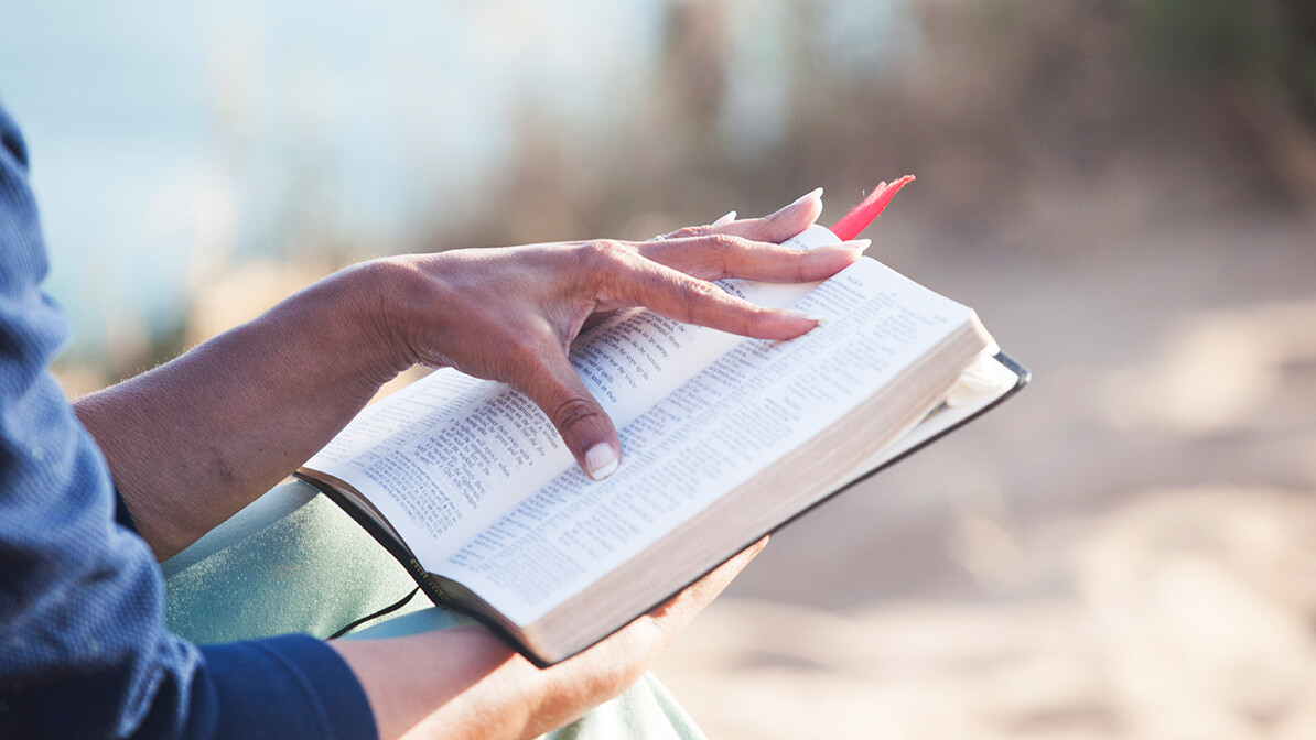 Hand resting on an open Bible outdoors in the sunlight.