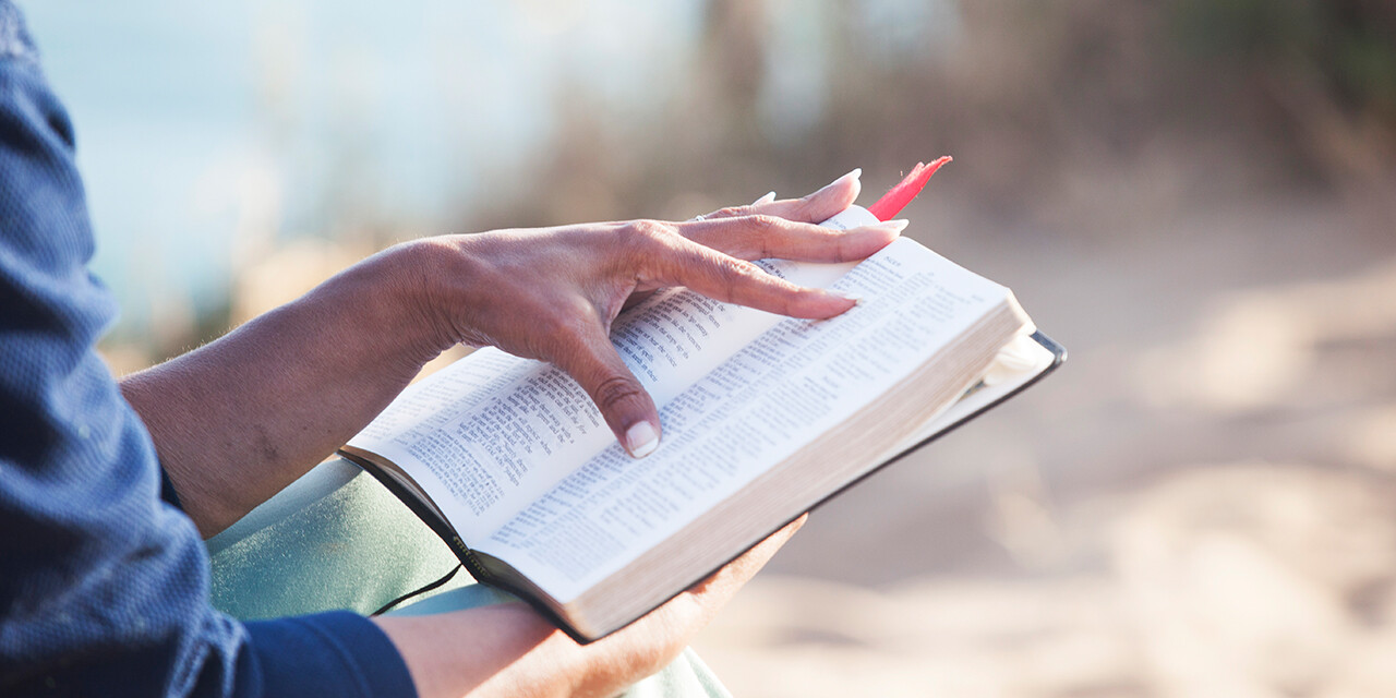 Hand resting on an open Bible outdoors in the sunlight.