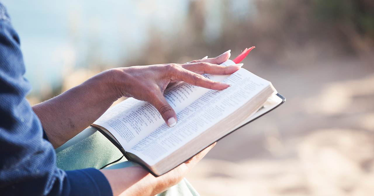 Hand resting on an open Bible outdoors in the sunlight.