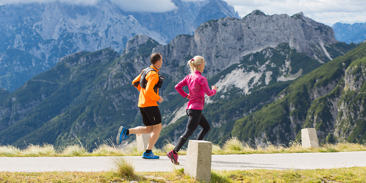 Man and woman running uphill on a trail beside mountains.