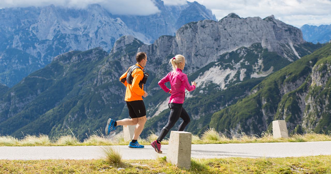 Man and woman running uphill on a trail beside mountains.