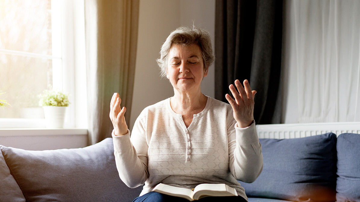 Older woman sitting in her living room with an open Bible on her lap and her hands raised.