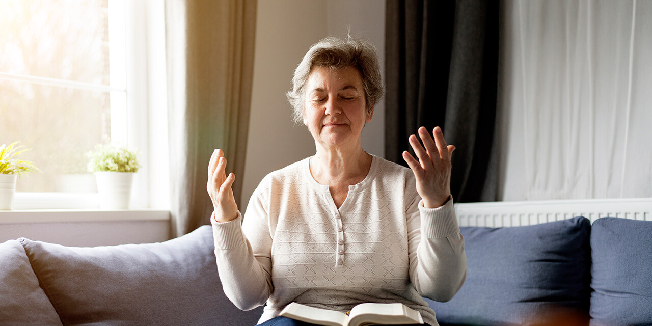 Older woman sitting in her living room with an open Bible on her lap and her hands raised.