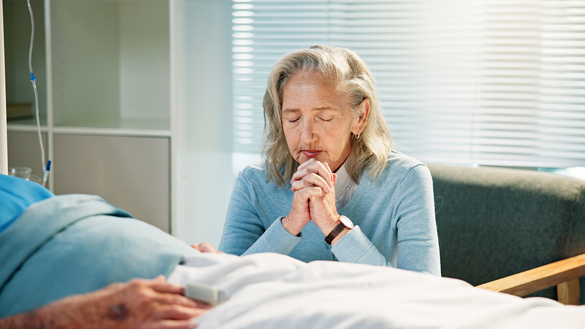 senior woman praying by the hospital bed of her husband, trusting God in financial hardship