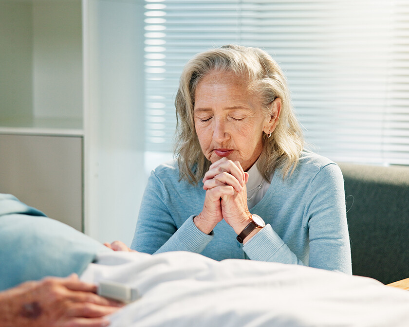 senior woman praying by the hospital bed of her husband, trusting God in financial hardship