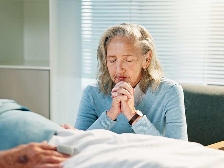 senior woman praying by the hospital bed of her husband, trusting God in financial hardship
